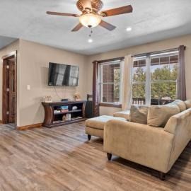 Living room with beige sectional, wall-mounted TV, ceiling fan, and large windows with brown curtains