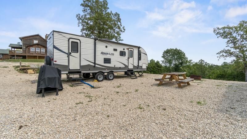 Camper trailer on gravel beside a picnic table under a cloudy sky, with trees in the background