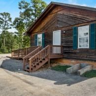 Wooden cabin with a front porch and stairs, set among pine trees beside a gravel driveway