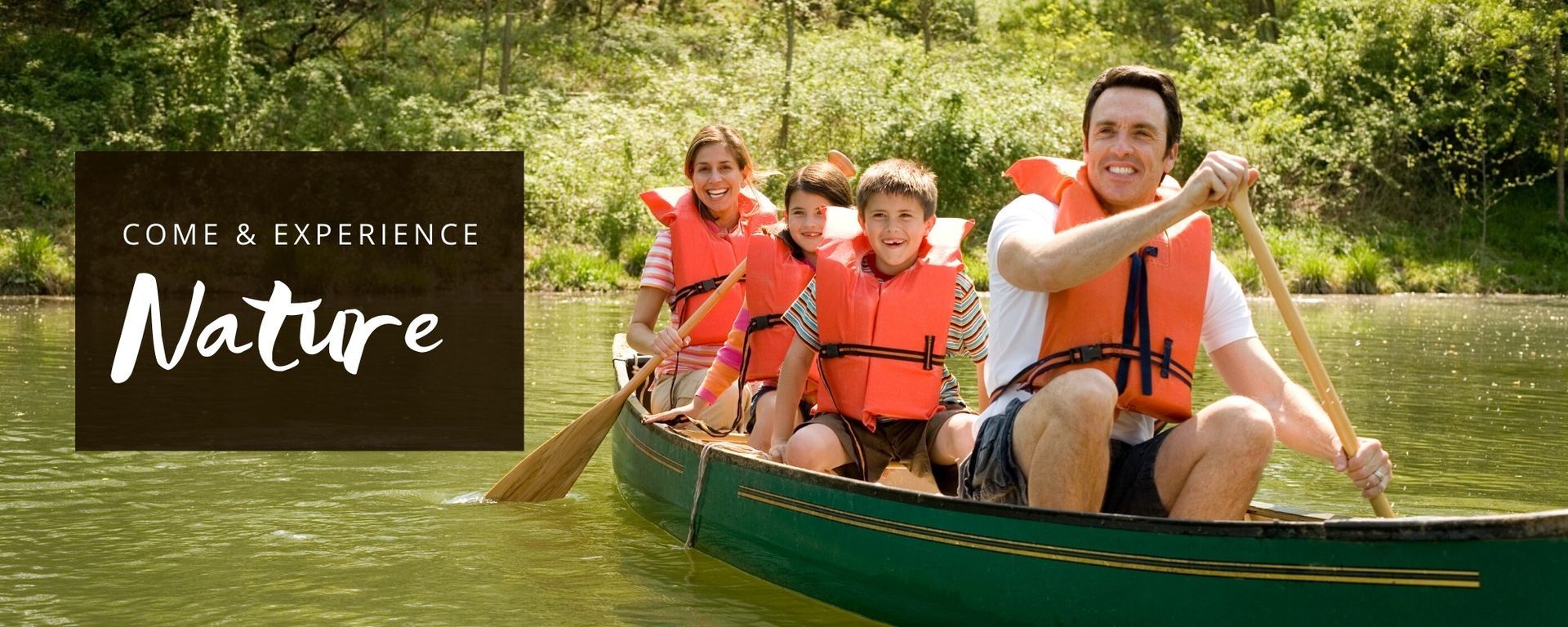 Family in orange life jackets paddles a green canoe on a calm lake beside a “Come & Experience Nature” sign.