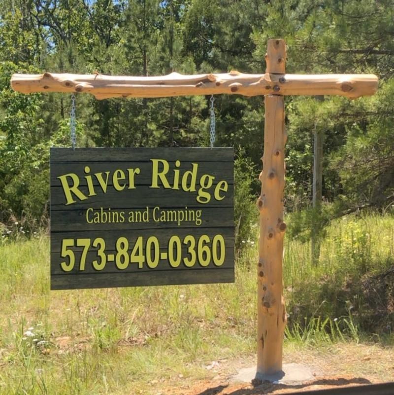 Rustic sign for River Ridge Cabins and Camping with phone number, hanging in a wooded area