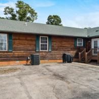 Single-story log cabin with gray roof, paved driveway, and wooden steps under a blue sky