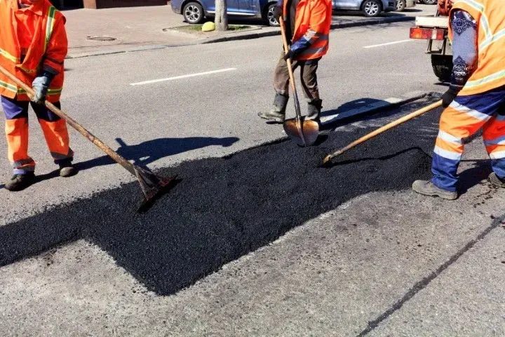 Three workers in high-visibility orange safety gear spread hot asphalt to patch a road surface.