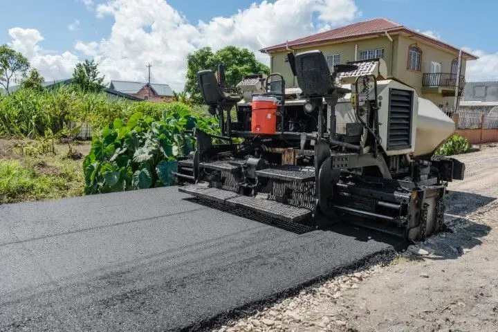 A road paver lays a fresh strip of asphalt on a dirt road in a sunny, residential neighborhood.
