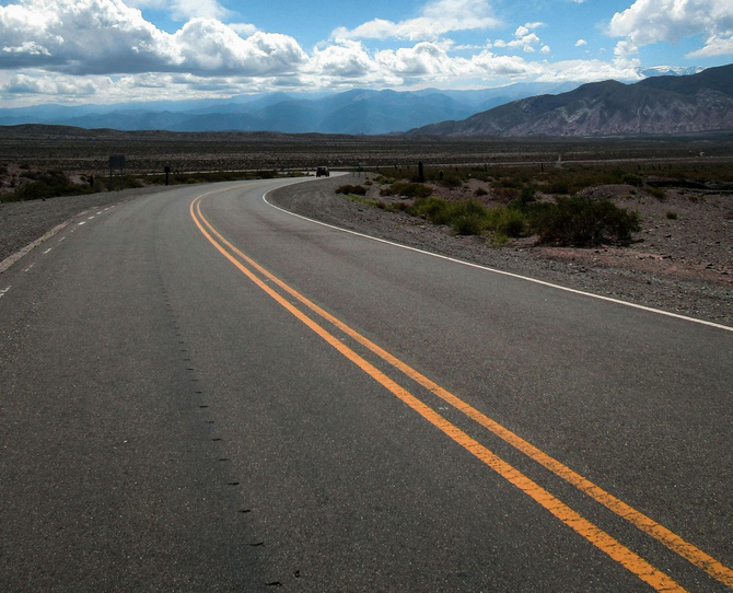 An asphalt road curves through a vast, arid landscape under a cloudy blue sky with distant mountains.