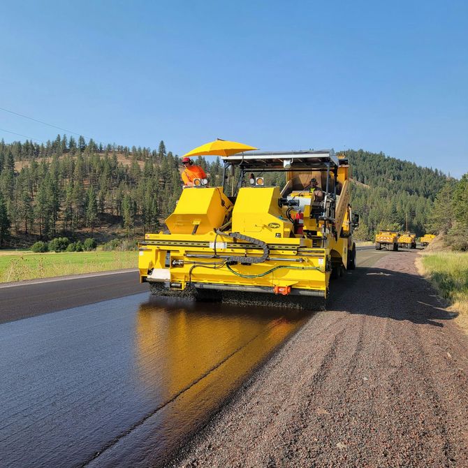 A yellow road construction machine applies a black seal coat to a gravel road surface on a sunny day.
