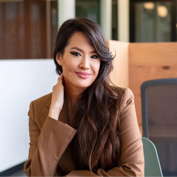 Smiling woman in a brown blazer seated indoors, resting her hand on her neck.