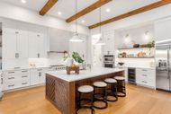 Modern kitchen with white cabinetry, a wood-paneled island with four bar stools, wood floors, and exposed ceiling beams.
