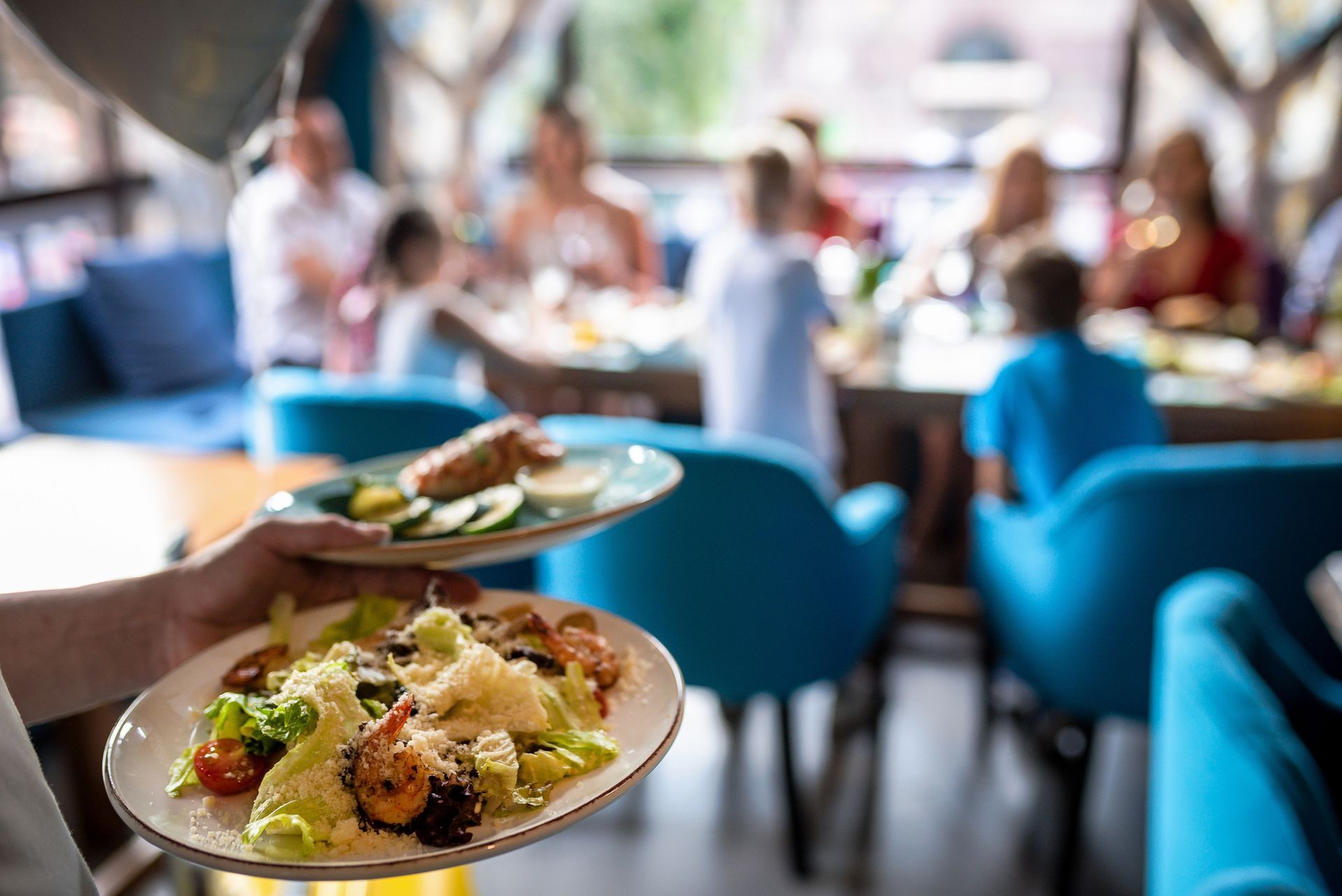 Cameriere che regge piatti di cibo mentre serve un gruppo di persone che cenano in un ristorante con sedie blu.