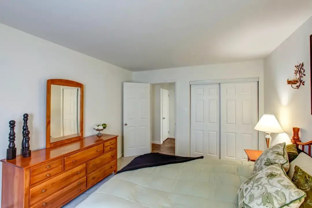 Bright bedroom with white walls, wood dresser, mirror, lamp, and bed with floral pillows