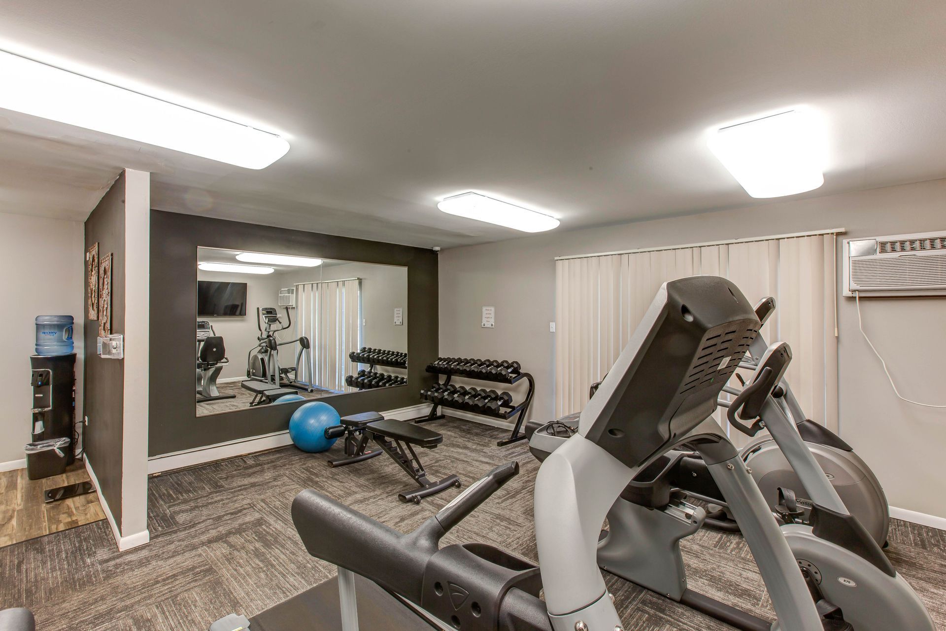 Home gym with treadmill, weight bench, dumbbells, mirror, and exercise ball under bright ceiling lights