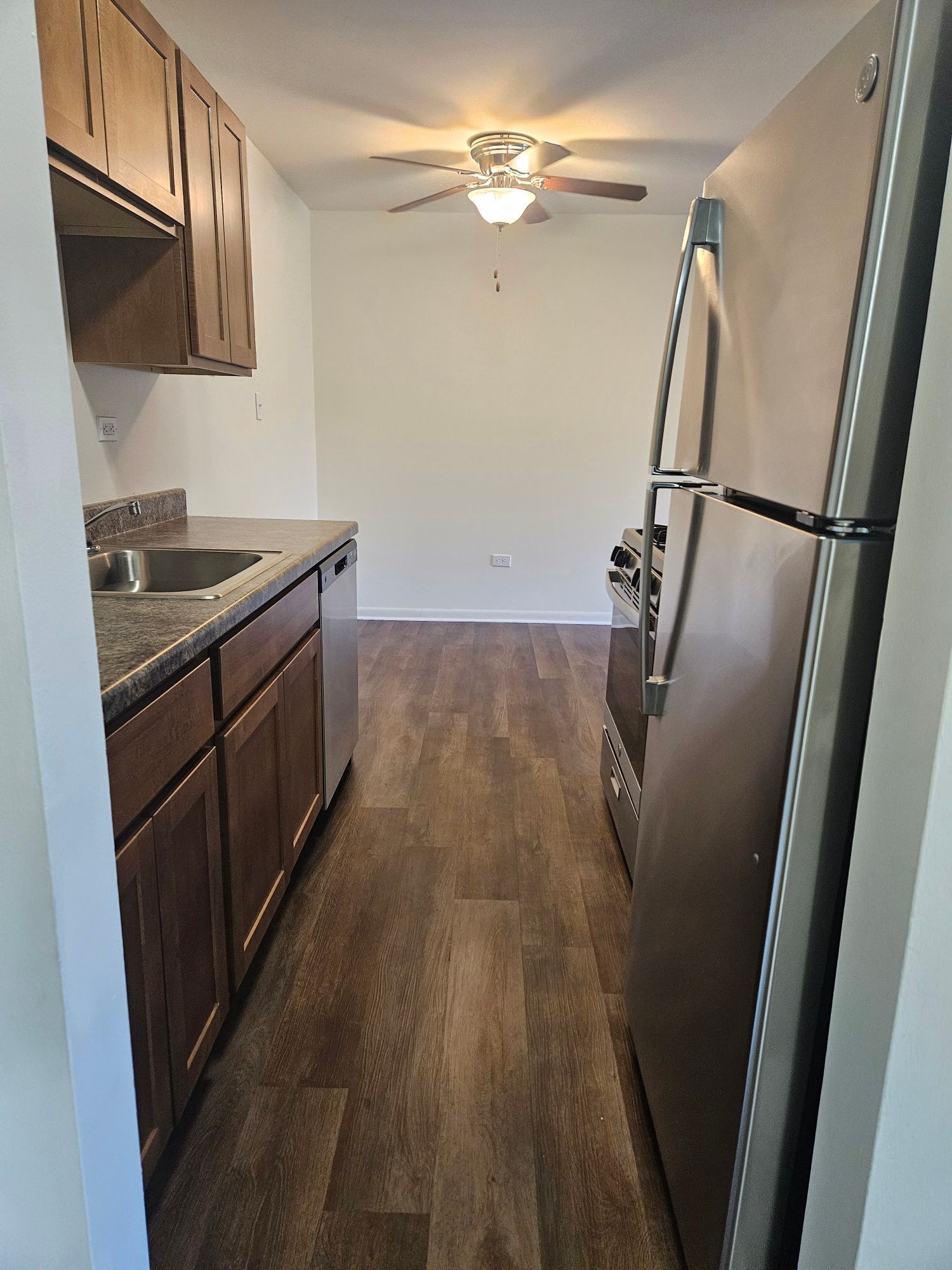 Narrow apartment kitchen with dark cabinets, granite counters, stainless fridge, and ceiling fan, leading to a room.