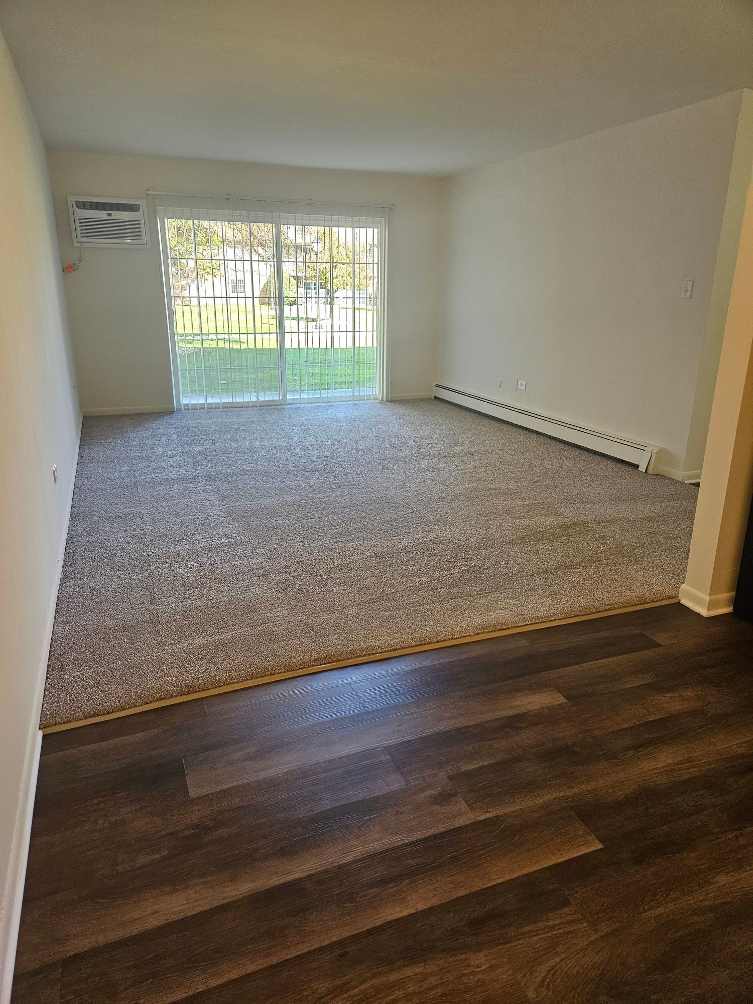 Empty carpeted living room with dark wood floor in foreground and sliding glass doors to a grassy patio.