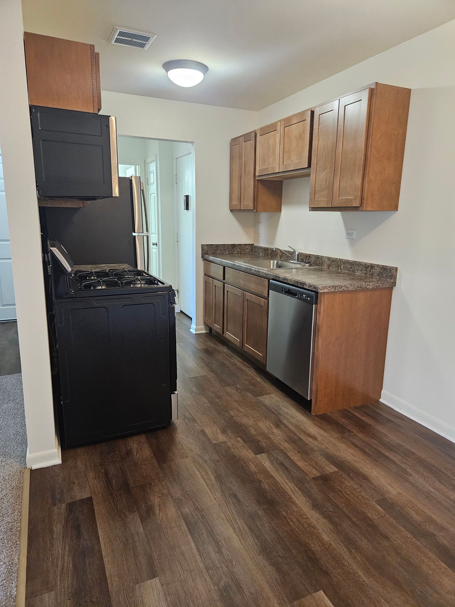Empty kitchen with wood cabinets, black appliances, granite counters, and wood-look flooring.