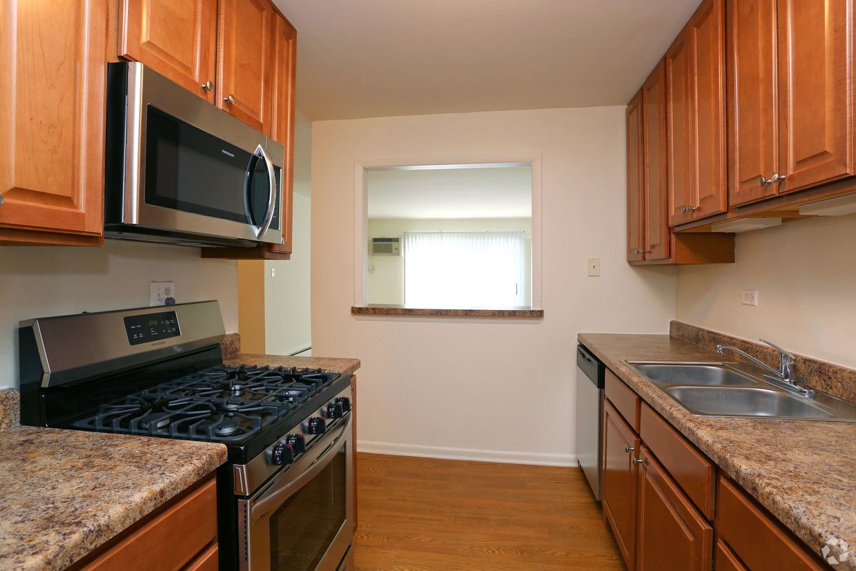Narrow galley kitchen with wooden cabinets, black stove, sink, and a small pass-through window.