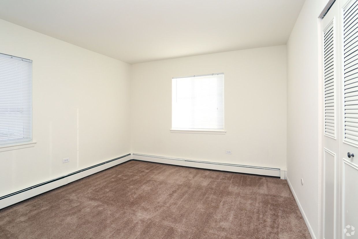 Empty carpeted room with white walls, windows, and closet doors.