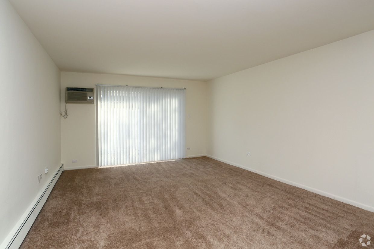 Empty carpeted living room with white walls and a sliding glass door with vertical blinds