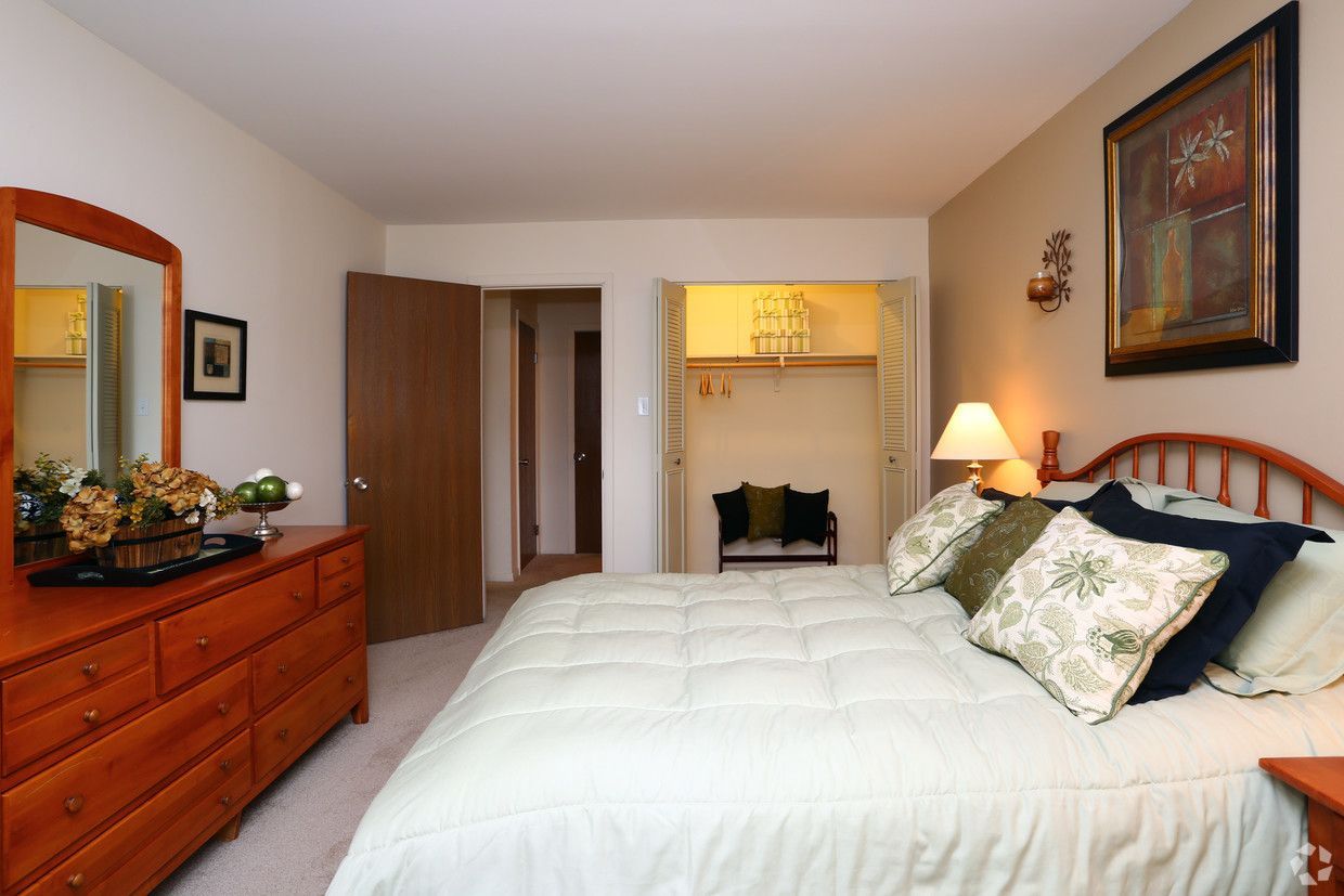 Bedroom with white bed, wooden dresser, warm lamp, and open closet in a tidy, softly lit room