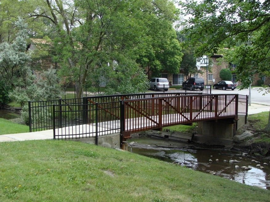 Small wooden footbridge over a creek in a green park, with trees and a sidewalk nearby