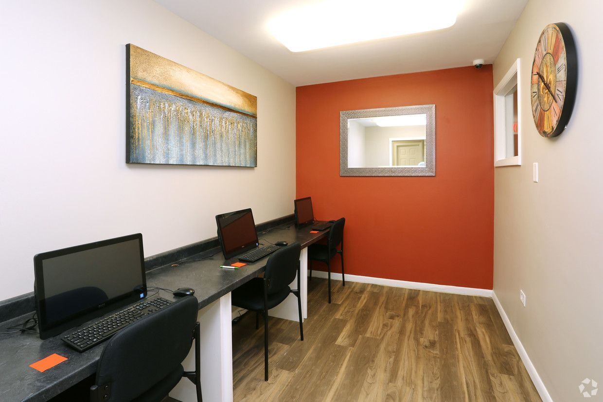 Modern office hallway with desks, laptops, wood flooring, and orange accent wall.