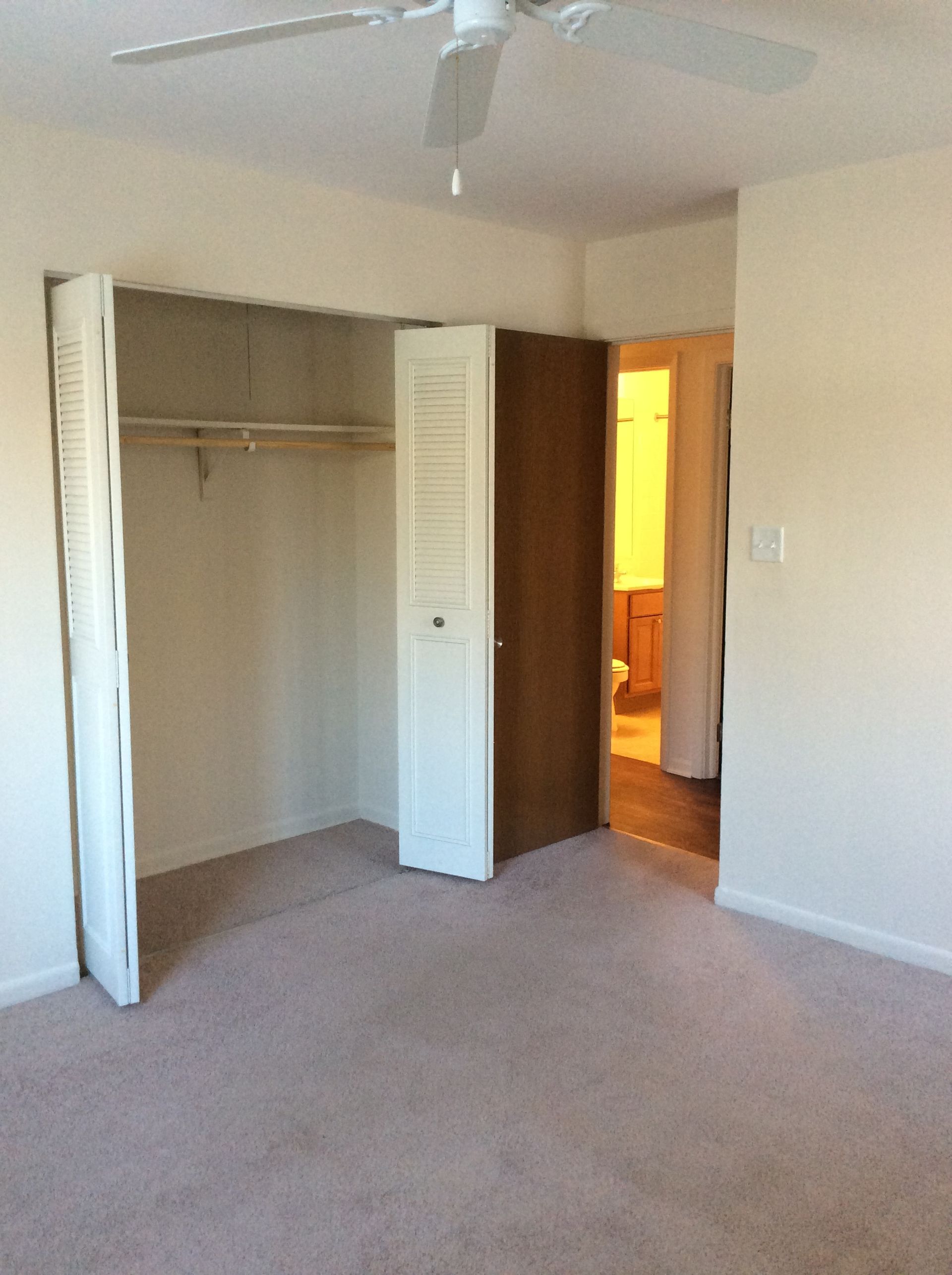 Empty beige bedroom with open closet doors and a doorway to a lit bathroom.