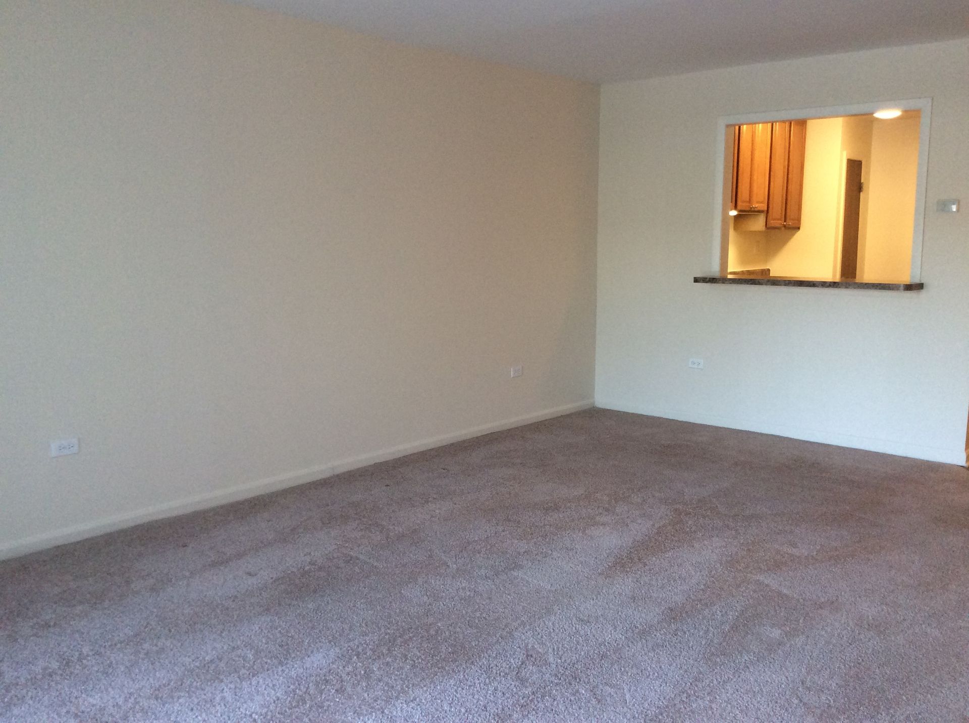 Empty beige carpeted living room with a pass-through opening to a kitchen
