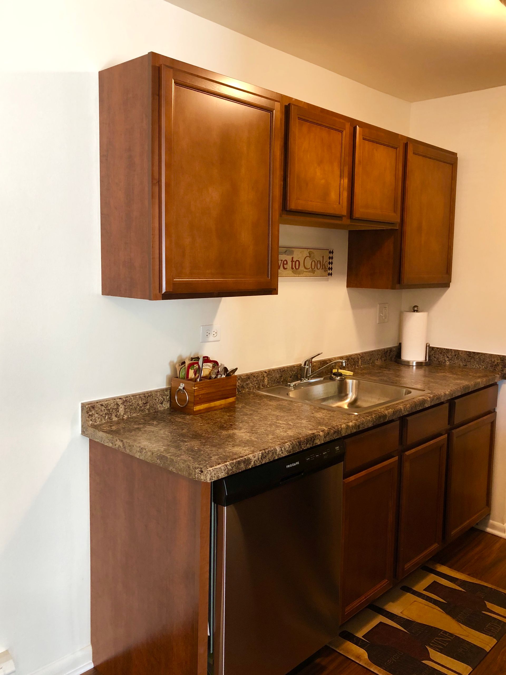 Small kitchen area with dark wood cabinets, granite countertop, sink, dishwasher, and patterned rug.