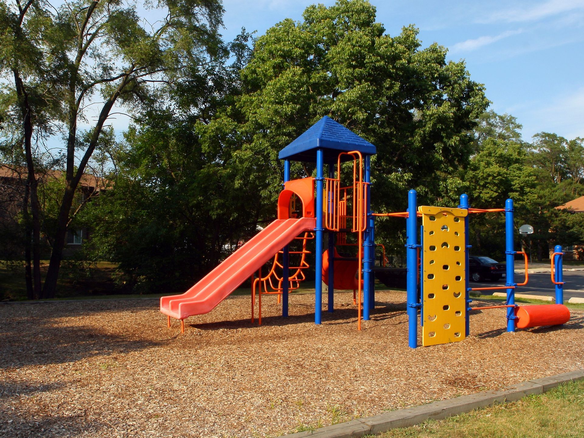 Colorful playground with red slide, climbing wall, and blue-orange play structures on wood chips under trees