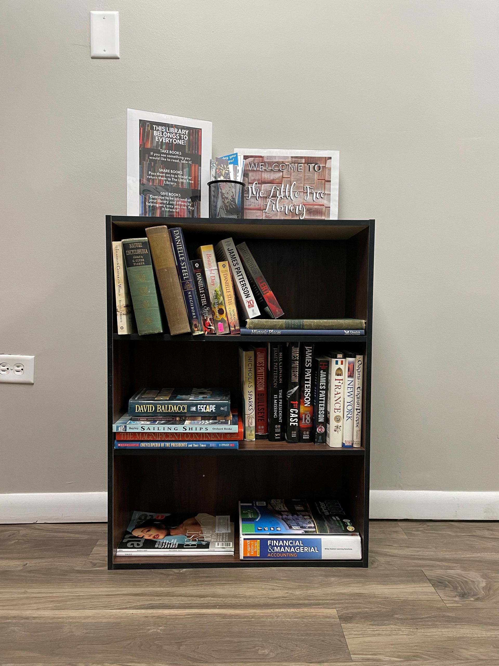 Small black bookshelf with books and board games against a gray wall on a wood floor.