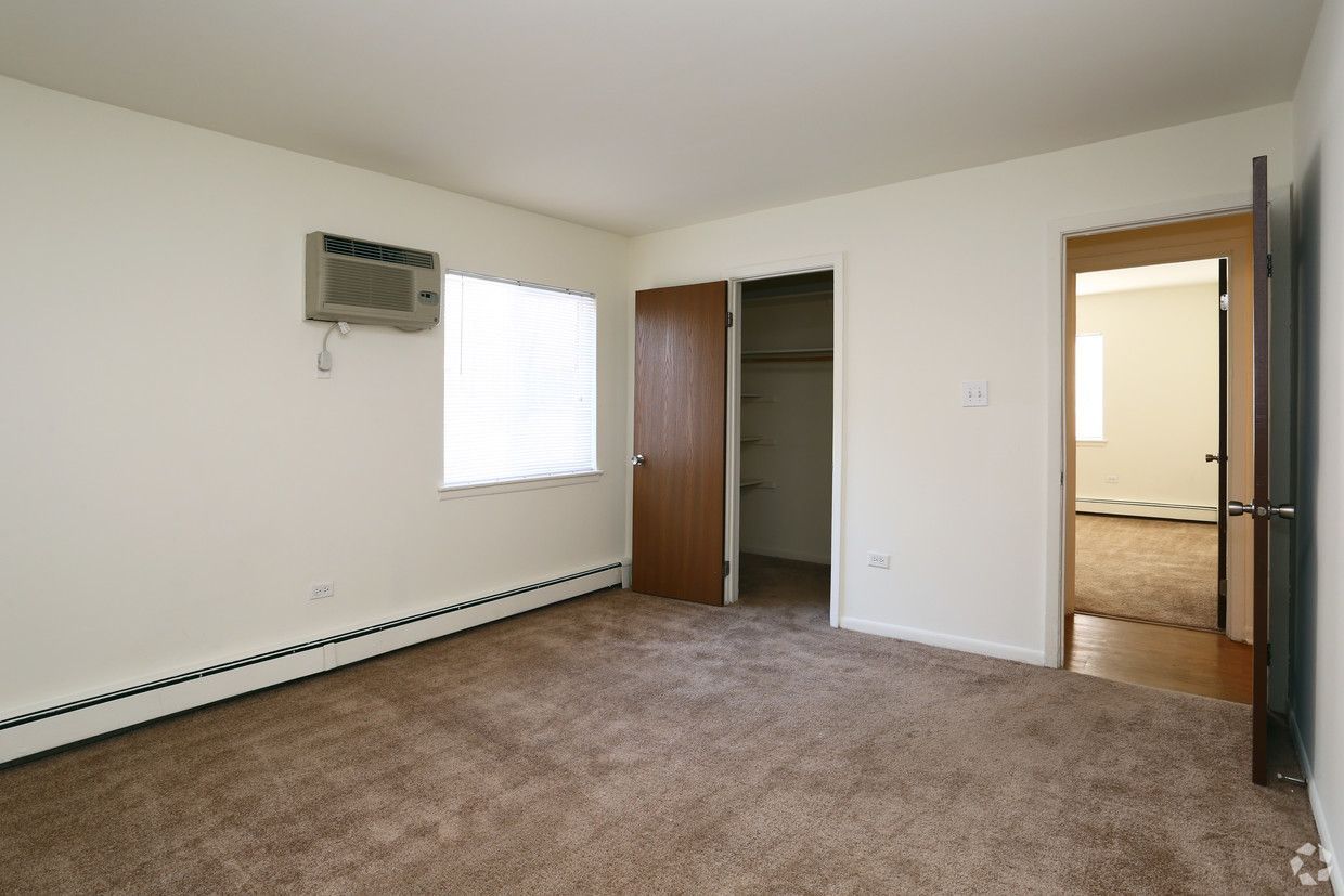 Empty carpeted room with wall-mounted AC, window blinds, and open doors to a hallway and bathroom.