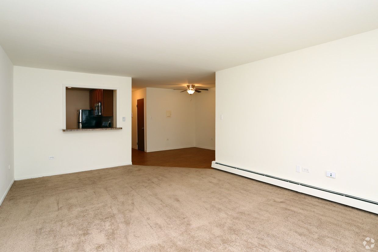 Empty carpeted living room with white walls and a pass-through opening to the kitchen.