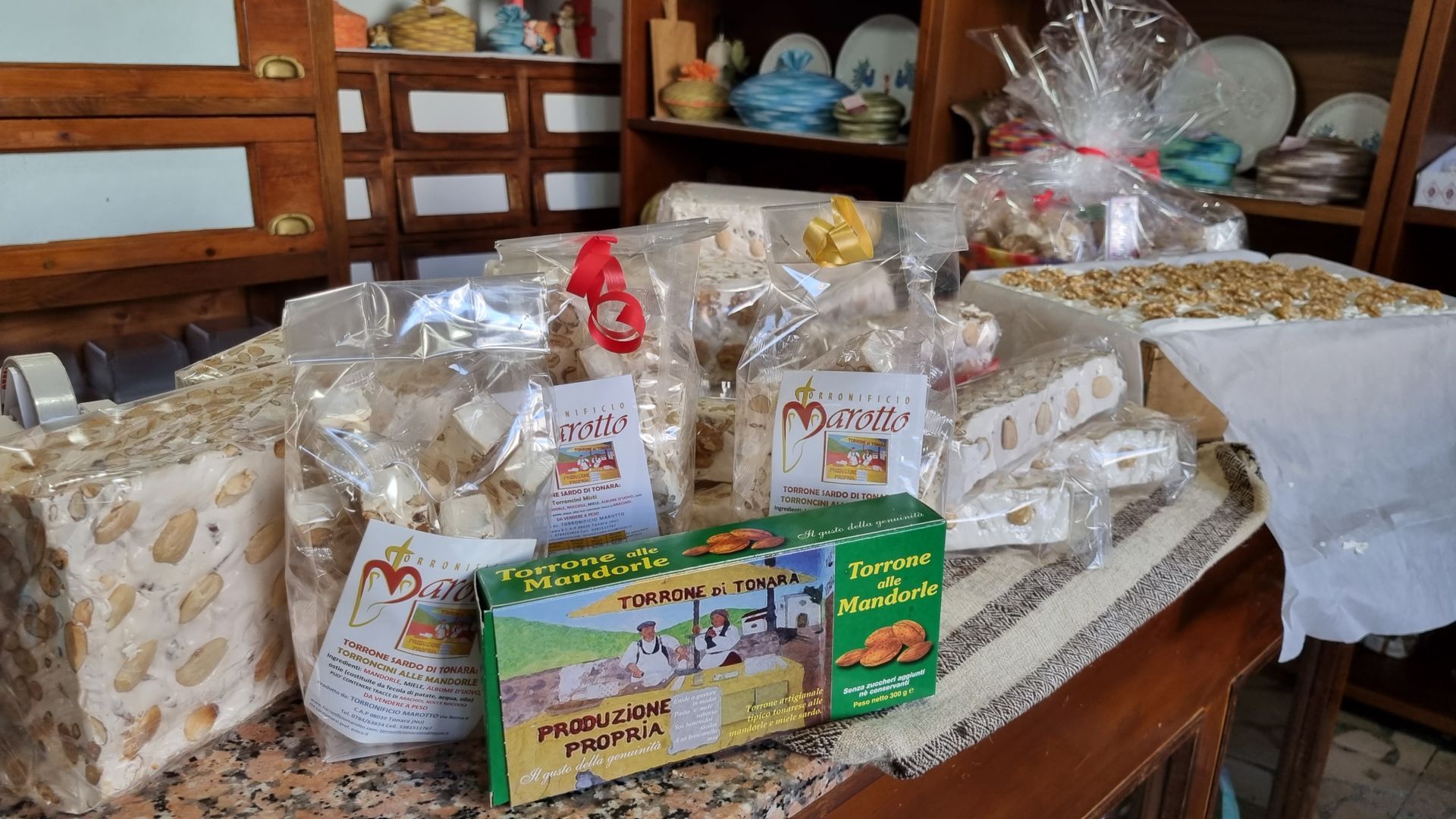 Assortimento di torrone confezionato e una scatola verde su un tavolo. Sullo sfondo, scaffali con oggetti decorativi.