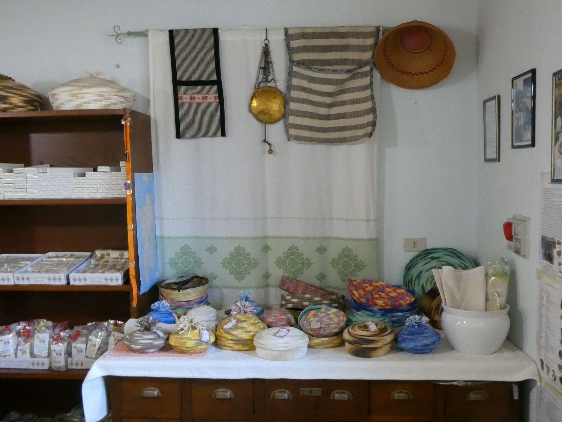 Esposizione di oggetti artigianali: cesti, tessuti e ceramiche su scaffali e un tavolo contro una parete bianca.