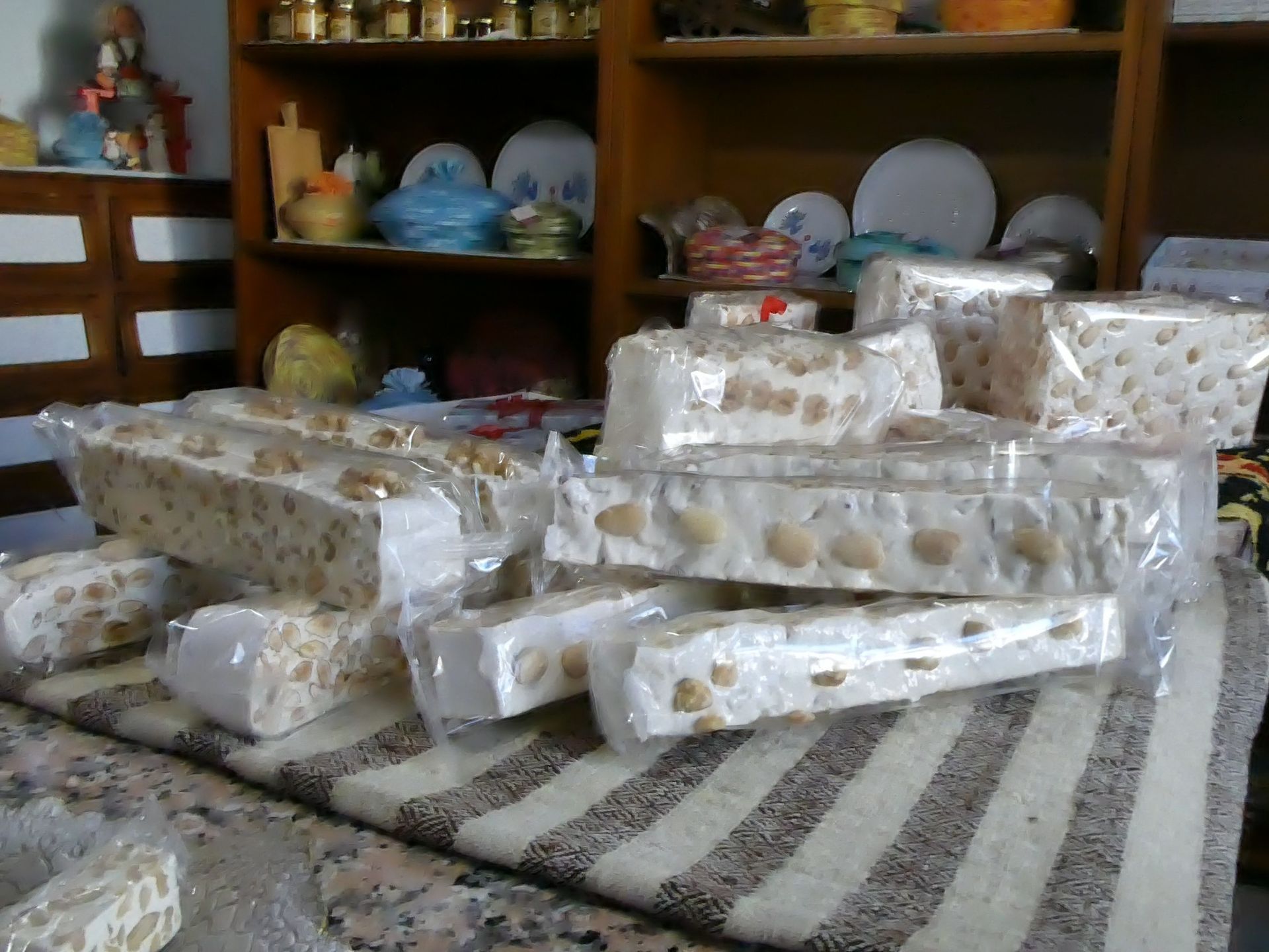 Su un tappeto a righe sono appoggiate pile di barrette di torrone, confezionate in plastica trasparente; sullo sfondo si trovano scaffali con oggetti decorativi.