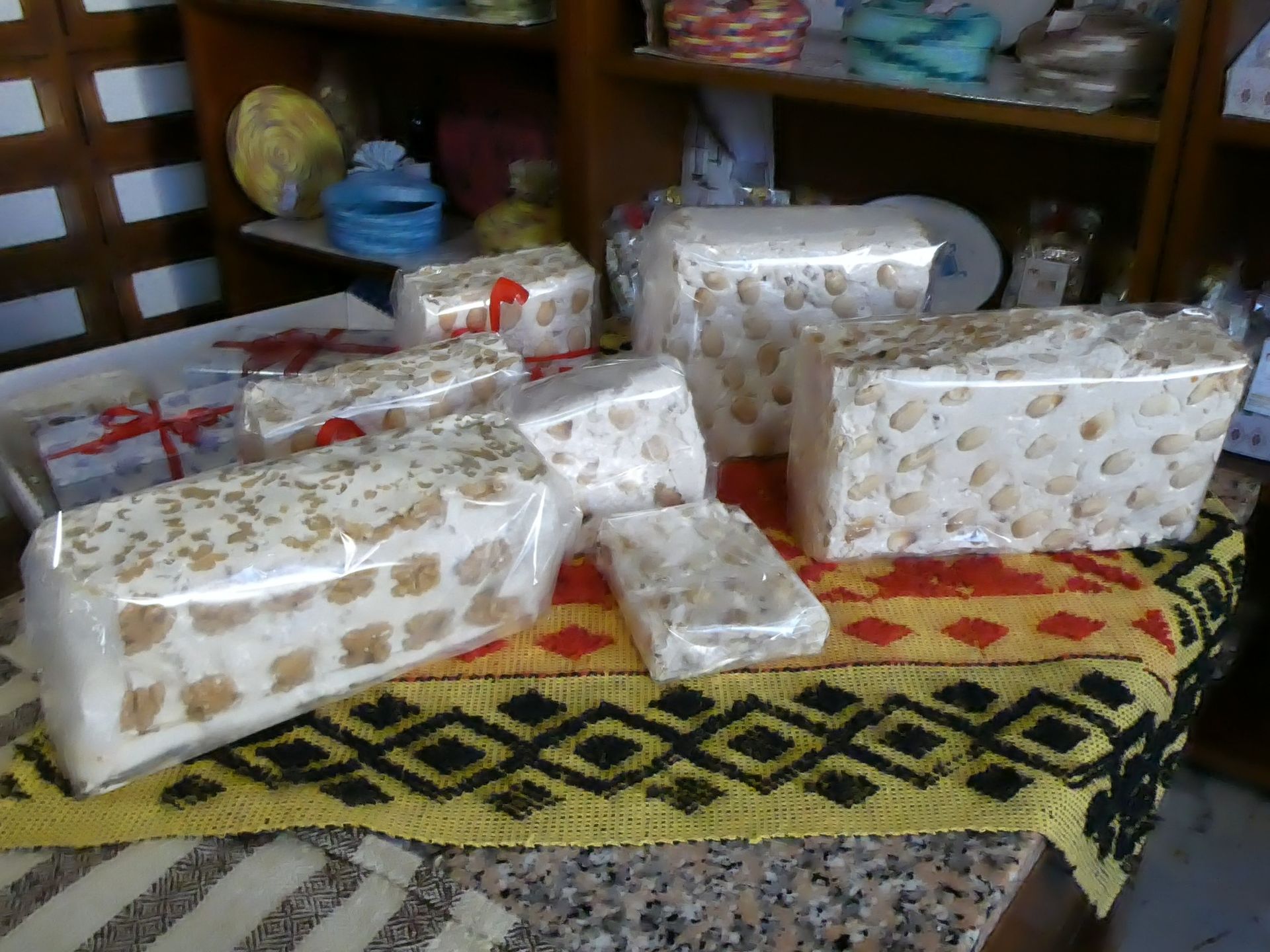 Blocchi di torrone avvolti in plastica trasparente, esposti su una tovaglia fantasia, con scaffali sullo sfondo.