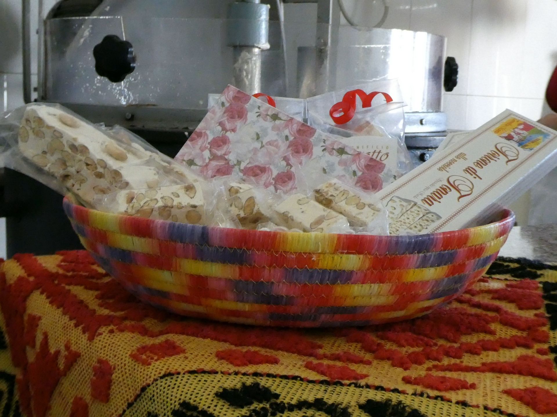 Cesto di torrone su un colorato cestino intrecciato, appoggiato su una tovaglia fantasia, con utensili da cucina sullo sfondo.