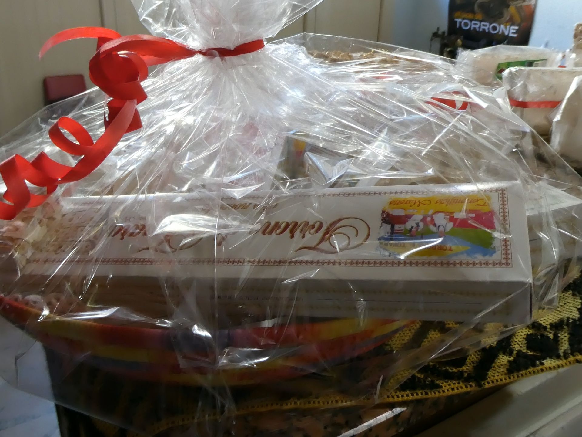 Cesto regalo avvolto in cellophane con un nastro rosso, contenente una lunga scatola di dolciumi.