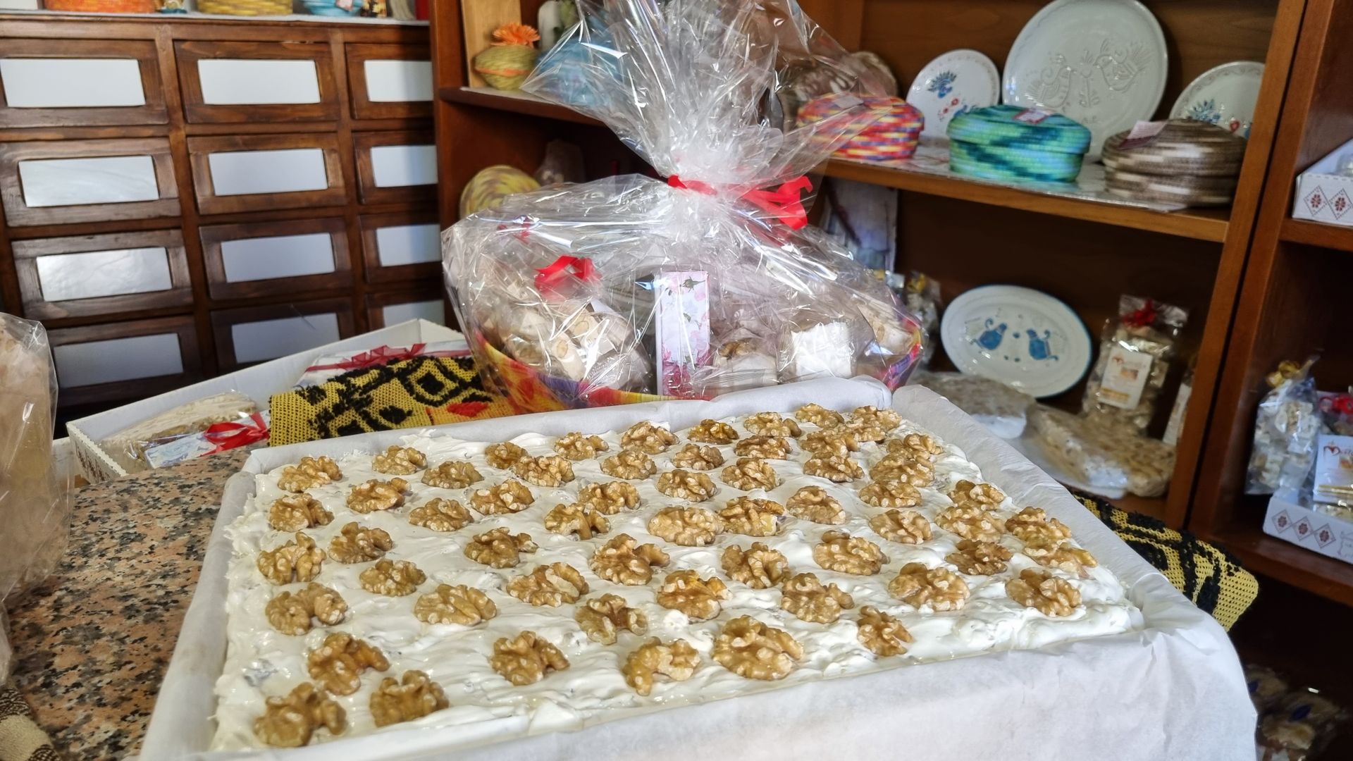 Un grande dolce rettangolare guarnito con noci è appoggiato su un tavolo. Dietro ci sono scaffali con vari articoli.
