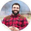 Man with beard smiles in red plaid shirt, brown collar detail, outdoor setting.