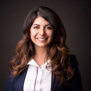 Woman with long wavy brown hair smiles at the camera, wearing a white shirt and navy blazer.