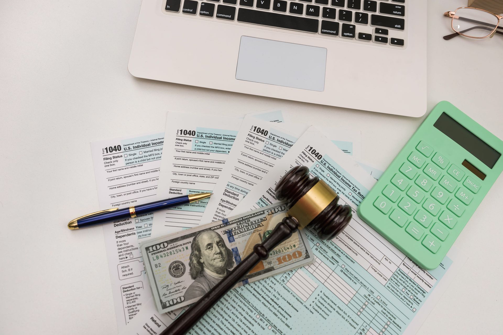 A laptop, tax forms, a stack of hundred-dollar bills, a wooden gavel, and a green calculator on a white desk.