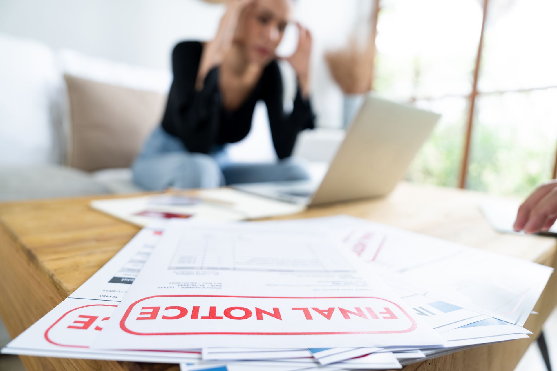Woman with head in hands, stressed, looking at bills, laptop in background. Final notice.