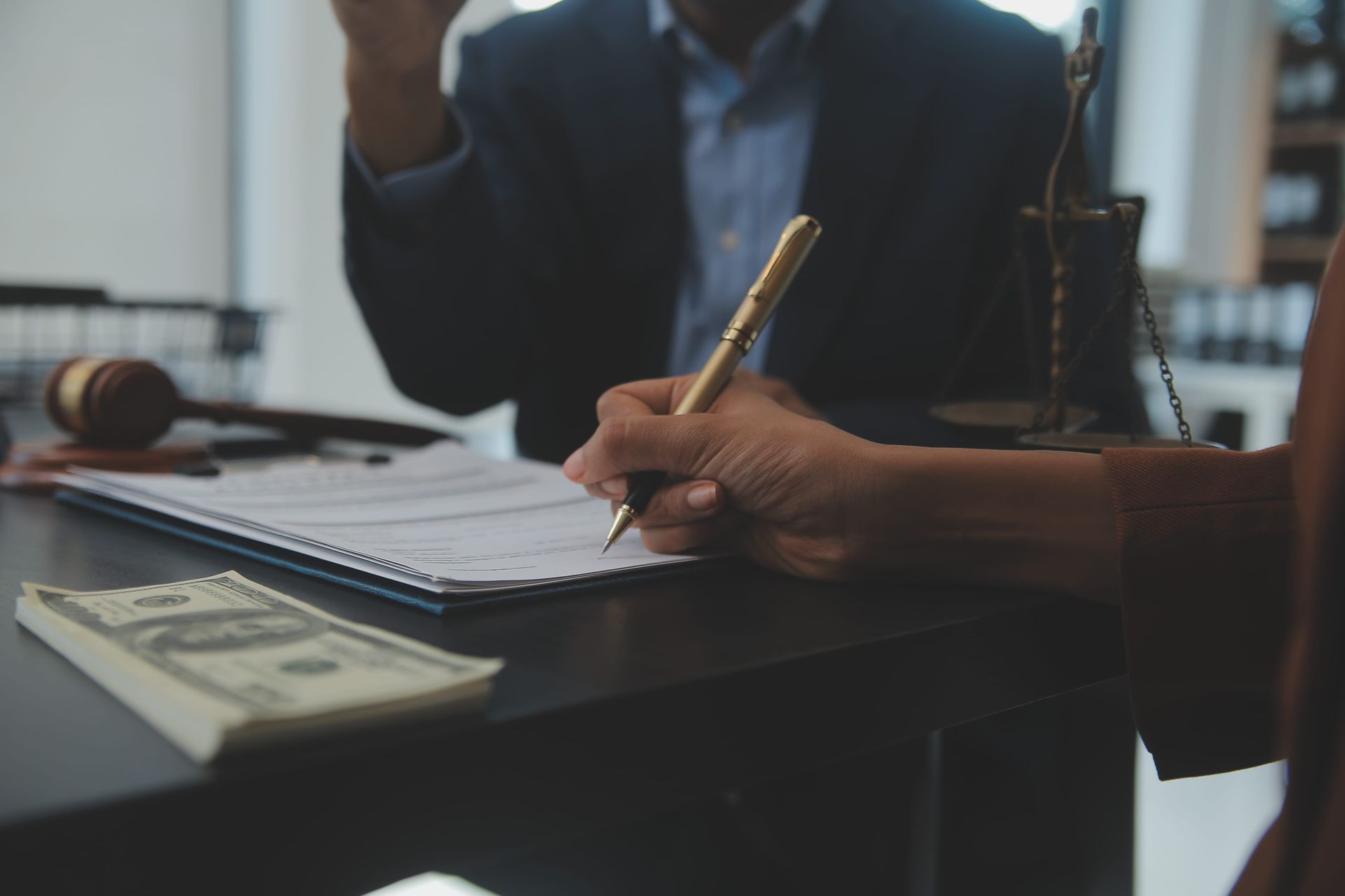 A person writing on a document in a law office with a gavel, legal scales, and a stack of cash on the table.