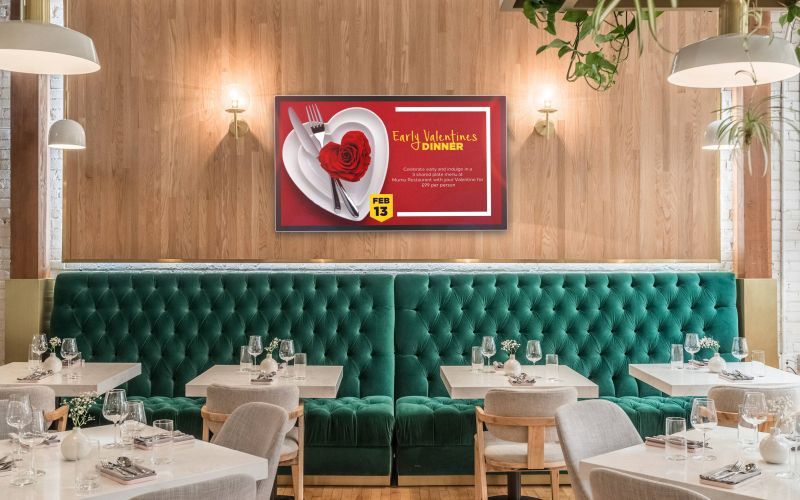 Restaurant interior with emerald booth seating, tables set with glassware, and a Valentine's Day promotion sign.