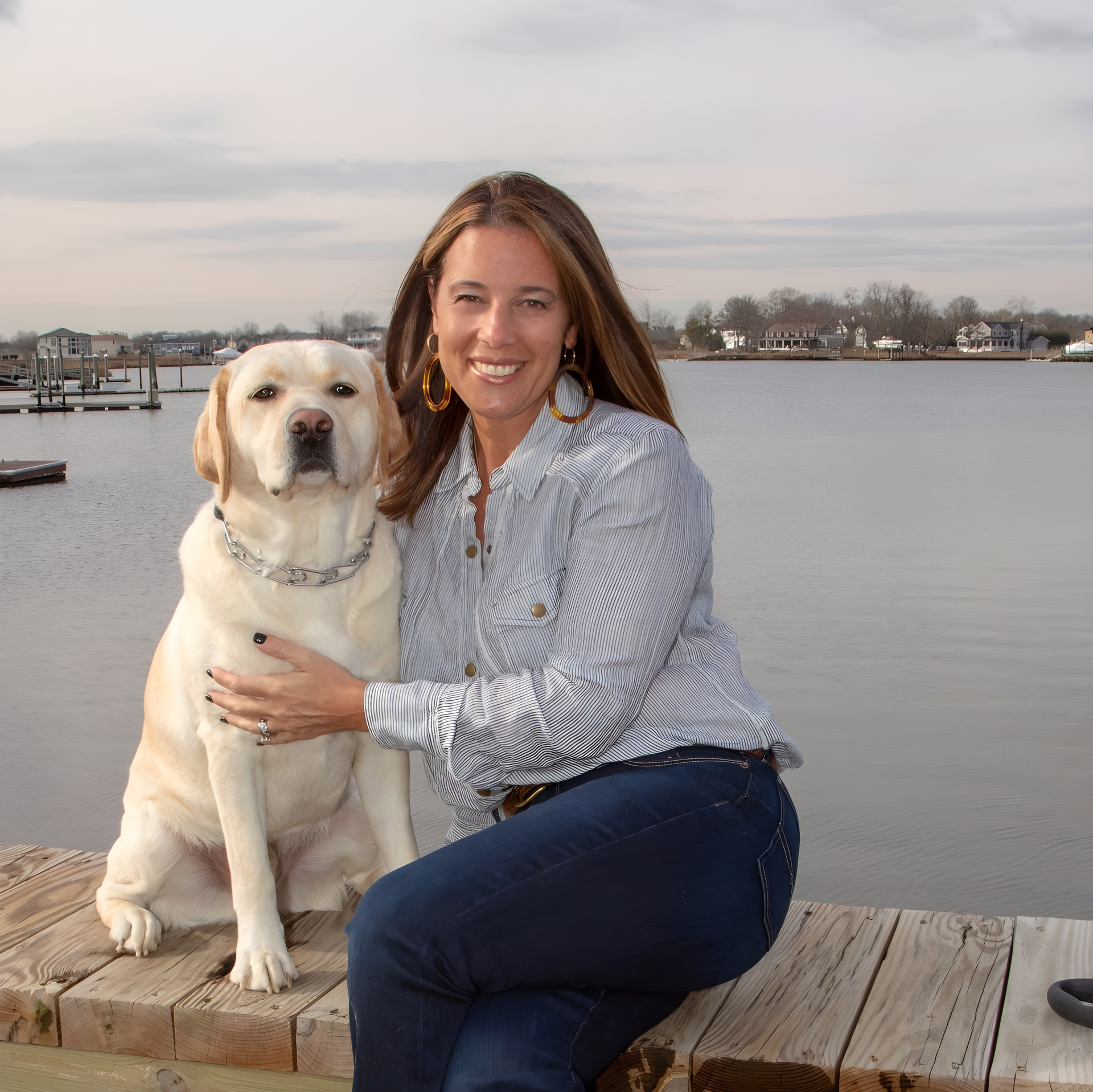 A smiling person sits on a wooden dock beside a large yellow Labrador retriever, with a calm bay in the background.