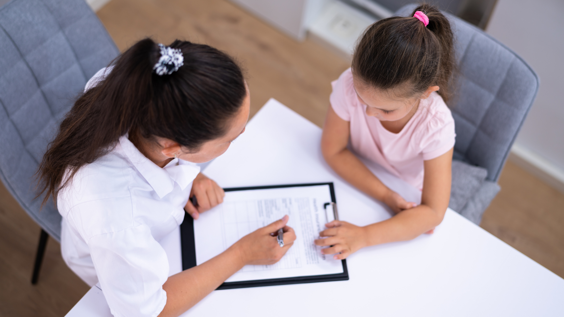A professional and a child seated at a white table, reviewing a document on a clipboard together.