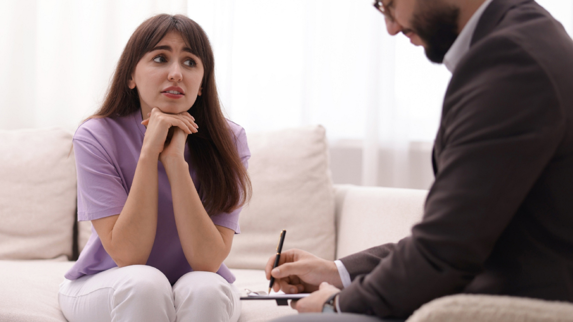 A person in a purple shirt sits on a sofa, looking toward a professional taking notes during a consultation.