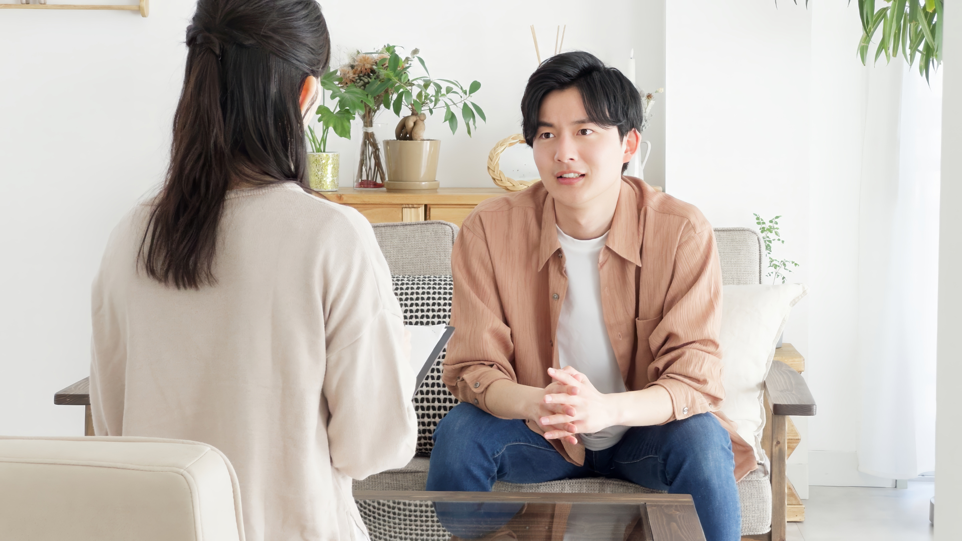 A person in a tan shirt sits on a sofa, speaking to someone with their back to the camera in a bright, indoor living space.