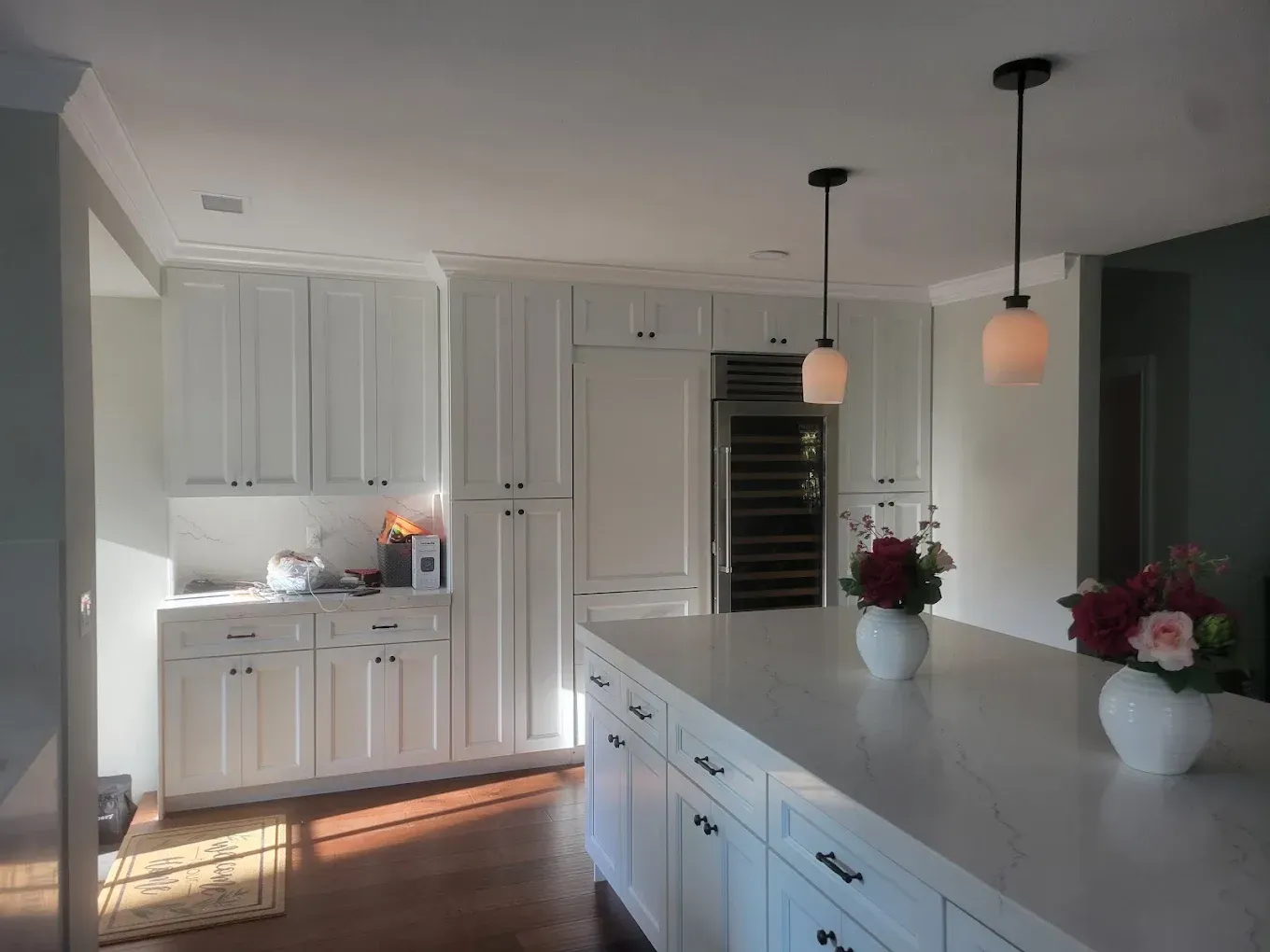 White kitchen with island, cabinets, wine fridge, and pendant lights; flowers on the island.