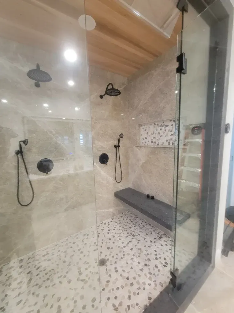 Modern bathroom shower with glass door, black fixtures, pebble floor, built-in bench, and tiled walls.